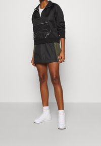 Svart zip-up sweatshirt med reflekterande logotyp, ihopparad med en mörkgrön och svart skort. Bärs med vita hög-top sneakers.