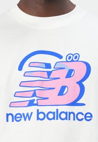 Hvid t-shirt med et stort multicolor "B" logo i pink og blå med bevægelseslinjer, samt "new balance" tekst i blå under logoet.