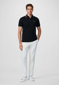 Camisa polo negra con acentos blancos en el collar y las mangas, combinada con pantalones blancos y zapatillas blancas, con un diseño ajustado.