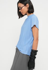 Ανοιχτό μπλε πλισέ μπλούζα με κοντά μανίκια, συνδυασμένη με μακριά γκρι φούστα και εφαρμοστά μαύρα δερμάτινα γάντια, που αναδεικνύουν μια λεία υφή.