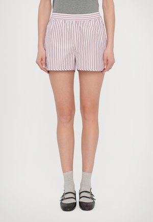 Pantaloni corti rosa chiaro con strisce rosse verticali, vita elastica, orlo scallop e tessuto leggero. Indossati con calzini grigi e scarpe nere.