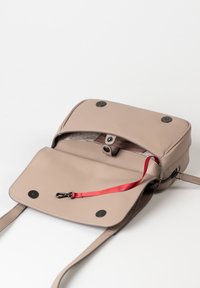 Beige Leder Umhängetasche mit einem Klappverschluss, zwei schwarzen Druckknöpfen und einem roten Innenriemen. Das Innere ist mit einem gemusterten Textil ausgekleidet.