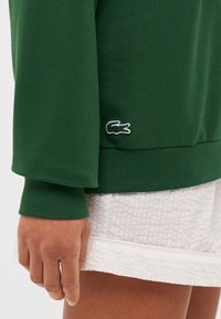 Groene katoenen sweatshirt met geribbelde manchetten, voorzien van een klein wit Lacoste krokodillenlogo aan de linkerkant, gecombineerd met gestructureerde witte shorts.
