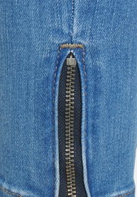 Modrá džínová tkanina s bočním zipem a metalickým povrchem, kontrastující se žlutým prošíváním podél švu.