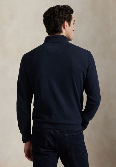 Homme de dos portant un pull à manches longues bleu marine foncé et un jean foncé sur un fond clair uni.