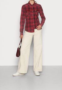 Camisa de cuadros roja y negra con dos bolsillos en el pecho, combinada con pantalones anchos de pana color crema. La modelo sostiene un pequeño bolso burdeos y lleva zapatillas blancas.