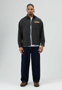 Zwart geruit overshirt met "FREEDOM" in geel geborduurd, gedragen over een wit T-shirt; gecombineerd met loszittende, donkerblauwe broek en bruine schoenen.