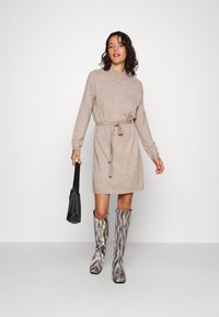 Robe en tricot beige avec un col haut et une ceinture à nouer, associée à des bottes hautes à motifs et un sac noir. Texture lisse.