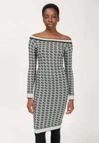 Frau in einem schwarz-weißen, schulterfreien Strickkleid mit einem sich wiederholenden geometrischen Muster, steht vor einem schlichten Hintergrund.