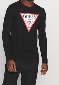 Mann trägt ein schwarzes Langarmshirt mit einem großen roten und weißen dreieckigen Guess-Logo auf der Brust, kombiniert mit schwarzen Hosen.