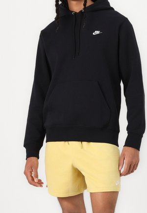 Mężczyzna ubrany w czarną bluzę z kapturem Nike z przednią kieszenią i żółte spodenki Nike, stojący na tle jednolitego białego tła.