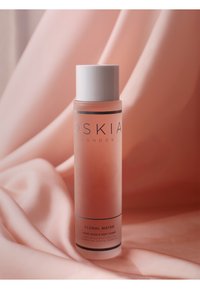 Transparent cylindrisk flaska med rosa blommvatten, vit kork, svart och silver etikett med texten "BLOMMVATTEN", vilande på mjuk rosa tyg.