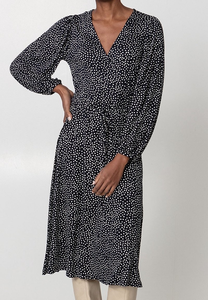 Femme portant une robe midi noire à pois blancs, manches longues, col en V, et ourlet à volants, debout avec une main sur la hanche.