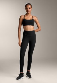 Črn športni modrček z tankimi naramnicami, črne hlače do pasu in črne atletske čevlje, ki se ponašajo z gladko teksturo tkanine.