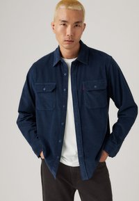 Marineblaues Hemd mit Knöpfen und zwei vorderen Taschen, aus strukturiertem Stoff, mit kragenartigem Ausschnitt und kontrastierenden Knöpfen, getragen über einem weißen T-Shirt.