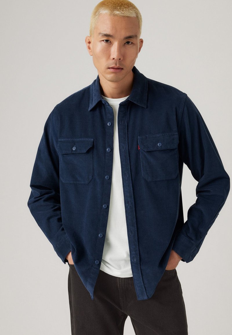 Marineblaues Hemd mit Knöpfen und zwei vorderen Taschen, aus strukturiertem Stoff, mit kragenartigem Ausschnitt und kontrastierenden Knöpfen, getragen über einem weißen T-Shirt.