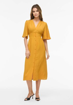 Femme portant une robe midi jaune moutarde boutonnée devant, manches aux coudes, et des sandales à talons noires à bout ouvert.