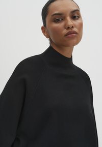 Maglione nero con collo alto e vestibilità rilassata, caratterizzato da una texture a coste sul colletto e sulle spalle, che mette in mostra un design minimalista.