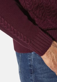Bordeauxroter Strickpullover mit strukturierten Zopfmustern an den Ärmeln und gerippten Bündchen, kombiniert mit blauen Denim-Jeans.