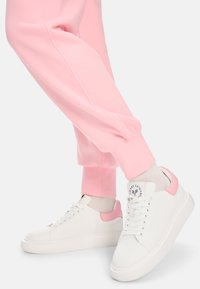 Weiße Sneaker mit einem pinkfarbenen Akzent an der Ferse, einer strukturierten Mesh-Oberfläche und flacher Sohle, kombiniert mit pinkfarbenen Jogginghosen am Knöchel.