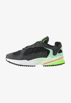 Scarpa atletica nera e grigia con materiali in rete e camoscio, accenti verdi, suola in gomma testurizzata e un design distintivo e aerodinamico.