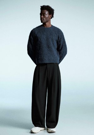 Marineblauw gebreide trui met een textuurafwerking, gecombineerd met zwarte geplooide broek en witte sneakers. Eenvoudige, moderne silhouet.