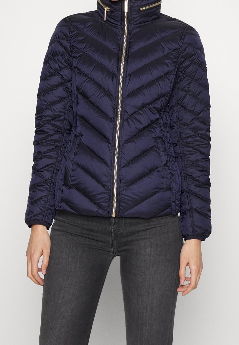 Granatowa kurtka puchowa w wzór zygzakowy, z zapięciem na zamek błyskawiczny, wysokim kołnierzem i marszczonymi rękawami. Posiada złote akcenty metalowe.