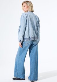 Femme blonde en veste en denim bleu clair et jeans évasés, se tenant tournée avec une main détendue, sur un fond blanc.