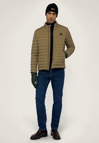 Zelená olivová vycpaná bunda s vertikálním šitím, vysokým límcem a zipem. Kombinovaná s černým svetrem, modrými džínami, zelenými rukavicemi a hnědými botami.