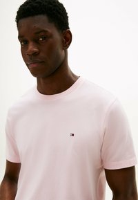 Jasnoróżowa koszulka T-shirt z bawełnianego materiału, z okrągłym dekoltem, z małym logo w czerwonym, białym i niebieskim kolorze po lewej stronie klatki piersiowej. Krótkie rękawy.