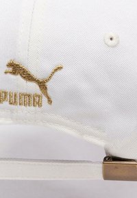 Gorra blanca de algodón con un acabado texturizado. Cuenta con un logo bordado en oro de un puma y texto, y con una hebilla metálica para el ajuste.