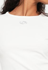 Biele tričko s okrúhlym výstrihom a krátkymi rukávmi, na prednej strane s malým strieborným logom. Mäkký bavlnený materiál s hladkou textúrou.