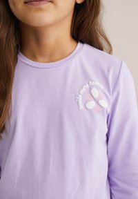 Fialkový top s dlhým rukávom s kvetinovým motívom s ružovými a bielymi okvetnými lístkami a textom "úsmev viac dnes" v bielom vyšívaní.