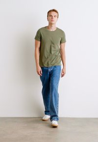 Olivgrünes T-Shirt mit kurzen Ärmeln, entspannter Schnitt. Kombiniert mit hellblauen geraden Jeans und weißen Sneakers, vor einem schlichten Wand-Hintergrund.