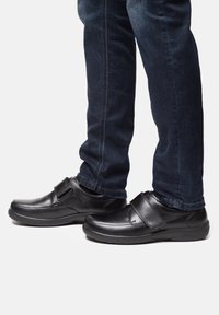 Scarpe nere in pelle con una cinghia in velcro, punta arrotondata e suola imbottita, abbinate a jeans blu scuro. Texture liscia e design semplice.