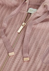 Rosa Zip-Hoodie mit gerippter Textur, ausgestattet mit einem goldenen Reißverschluss und passenden Kordelzügen. Etikett sichtbar am Halsbereich.