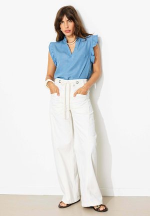 Femme aux cheveux bruns portant un chemisier bleu sans manches, un pantalon blanc taille haute à jambes larges et des sandales, debout les mains dans les poches.