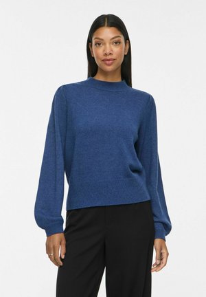 Femme aux longs cheveux noirs portant un pull bleu à manches longues et un pantalon noir, debout devant un fond blanc uni.