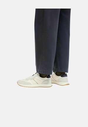 Persona che indossa scarpe da ginnastica bianche con accenti beige e neri, abbinate a pantaloni ampi blu scuro su uno sfondo semplice.