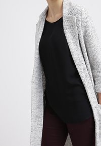 Harmaa teksturoitu pitkä takki, jossa on avoin etuosa, pukeutuneena mustan paidan ja tumman burgundin värisiin housuihin. Huomattava kevyestä kankaastaan ja rentosta istuvuudestaan.