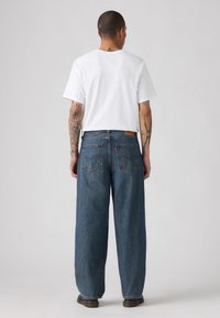 Hombre con tatuajes, vistiendo una camiseta blanca sencilla y unos pantalones vaqueros azules anchos, de pie de espaldas contra un fondo neutro.