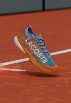 Zapatilla de running con diseño azul y naranja, gran marca "LACOSTE", volando sobre una pista de tenis de arcilla con partículas de polvo rojo.