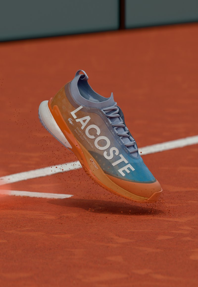 Chaussure de course au design bleu et orange, avec un grand logo "LACOSTE", en train de voler au-dessus d'un court de tennis en terre battue, des particules de poussière rouge s'en échappant.