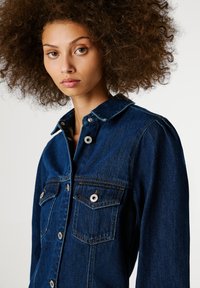 Tumši zila džinsa jaka ar pogu aizdari, divām krūšu kabatām un kniedēšanas aizdari. Iezīmēts ar norādītu apkakli un redzamām šuvju detaļām.