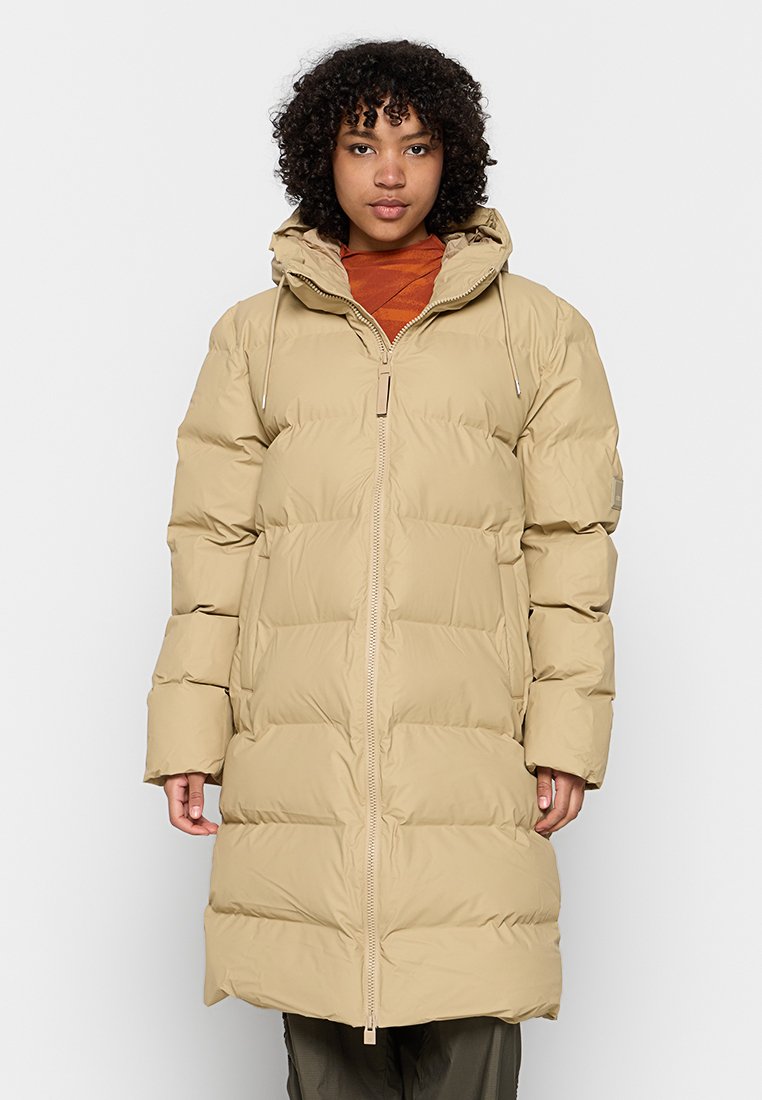 Manteau en duvet beige avec un design long et surdimensionné, une fermeture éclair à l'avant et une capuche. Présente une texture matelassée et des cordons de serrage ajustables.