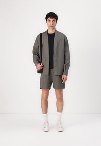 Giacca khaki a maniche corte con due tasche, abbinata a pantaloni corti coordinati. Il modello indossa una T-shirt nera e sneaker bianche con calze corte.