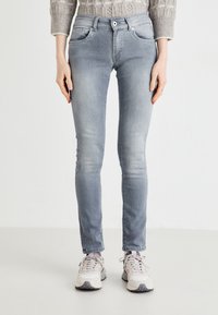 Jean skinny gris clair avec une texture lisse, poches avant et fermeture à bouton. Porté avec des baskets claires, sur un fond neutre.
