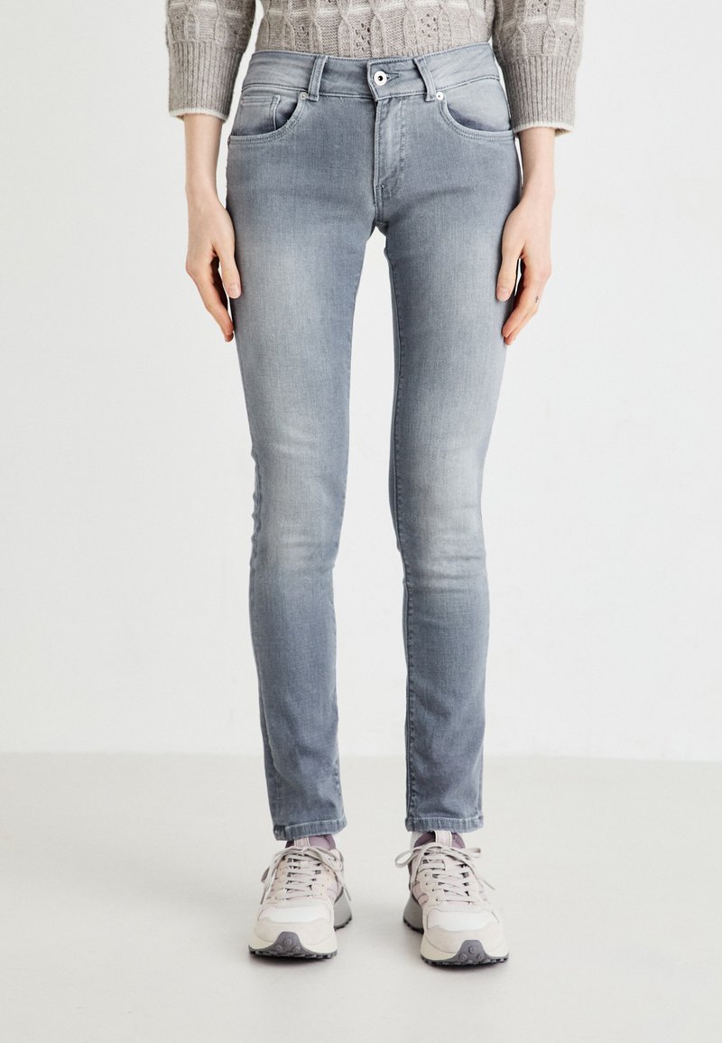 Jean skinny gris clair avec une texture lisse, poches avant et fermeture à bouton. Porté avec des baskets claires, sur un fond neutre.