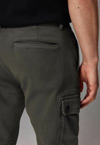 Dunkelgrüne Cargo-Hosen verfügen über eine Seitentasche mit Knopfverschluss und eine Gesäßtasche. Das Material scheint weich mit einem leichten Glanz zu sein.
