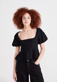 Top nero strutturato con scollatura quadrata e maniche a sbuffo, caratterizzato da un design a peplo e un delicato motivo a onde. Indossato con pantaloni scuri.